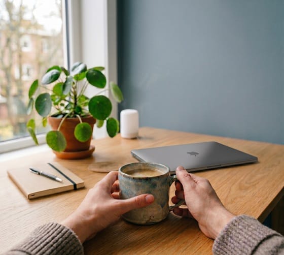 Un utilisateur qui tient une tasse de café devant un cahier de note et un portable fermé.